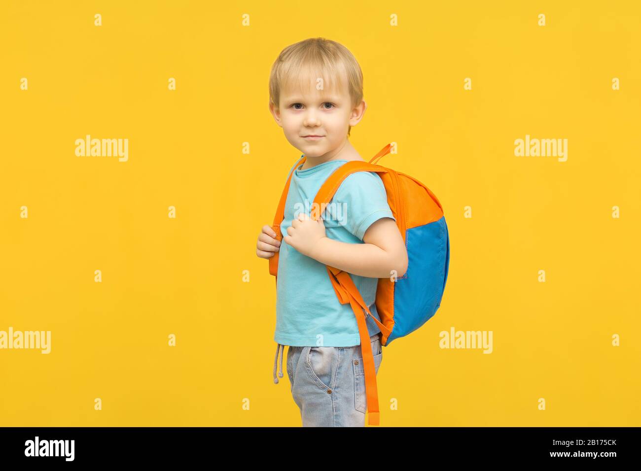 Freudiges Knabenkind steht mit einem Rucksack auf dem Rücken und blickt auf einem leuchtend gelben Hintergrund auf die Kamera. Idee für Reiseartikel und bequeme BA Stockfoto