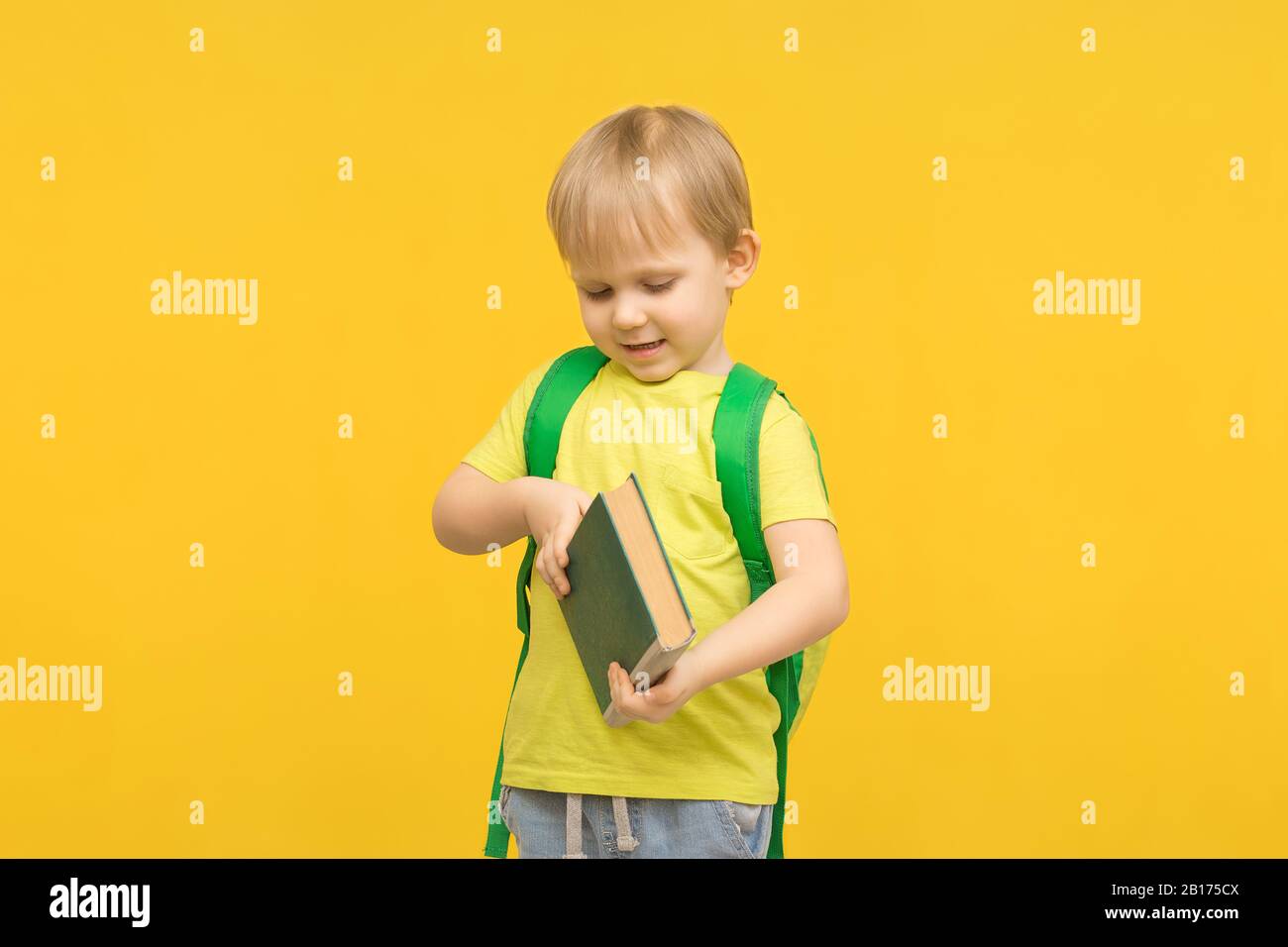 Kinderblonder Junge mit Rucksack öffnet ein Buch auf einem gelb hellen Hintergrund. Konzept für Artikel über Studium, Lesen, Kindheit und Vorschule Childre Stockfoto