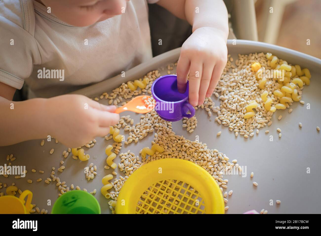 Kleines Kind gießt den gelben Löffel des Spielzeugs voll in der Tasse. Spiele mit Produkten. Montessori-Technik: Entwicklung von guten motorischen Fähigkeiten bei Kindern Stockfoto