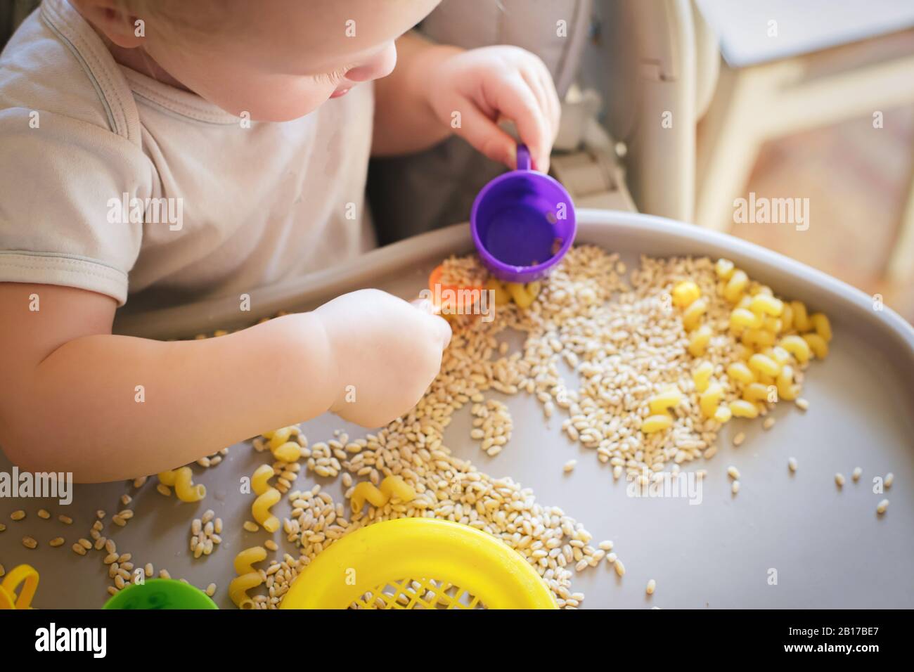 Kleines Kind gießt den gelben Löffel des Spielzeugs voll in der Tasse. Spiele mit Produkten. Walfdorfs Methode der Kinderentwicklung. Kinder lernen, selbstständig zu essen. Deve Stockfoto