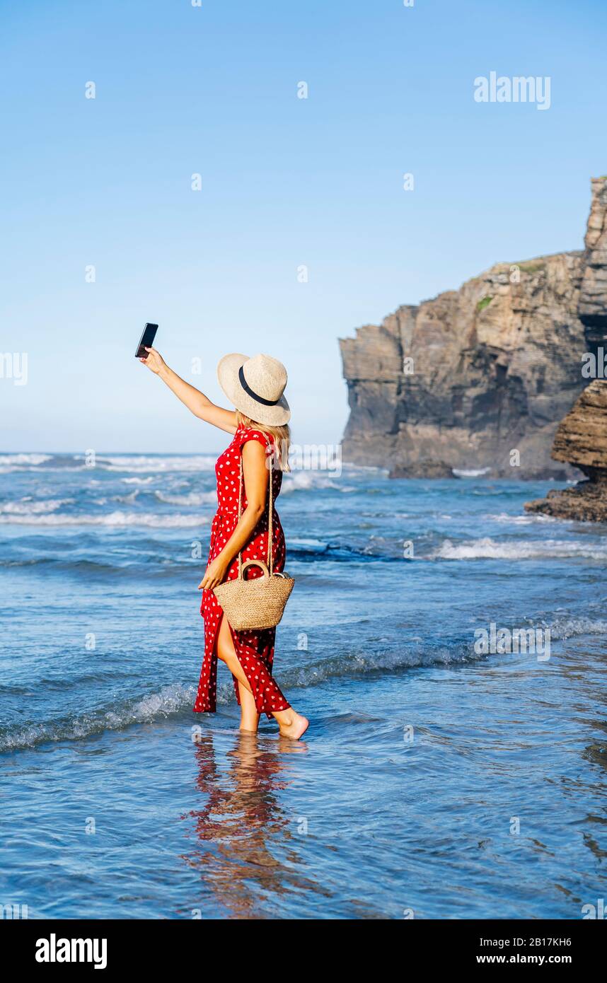 Blonde Frau mit rotem Kleid und Hut und Smartphone am Strand Stockfoto