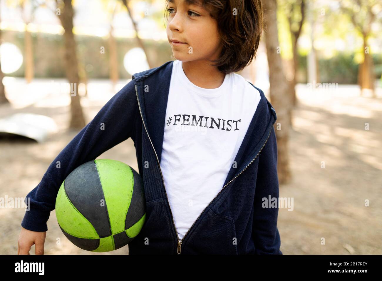 Kleiner Junge, der auf der Straße mit Aufdruck auf dem T-Shirt steht, sagt Feministin, hält Ball Stockfoto