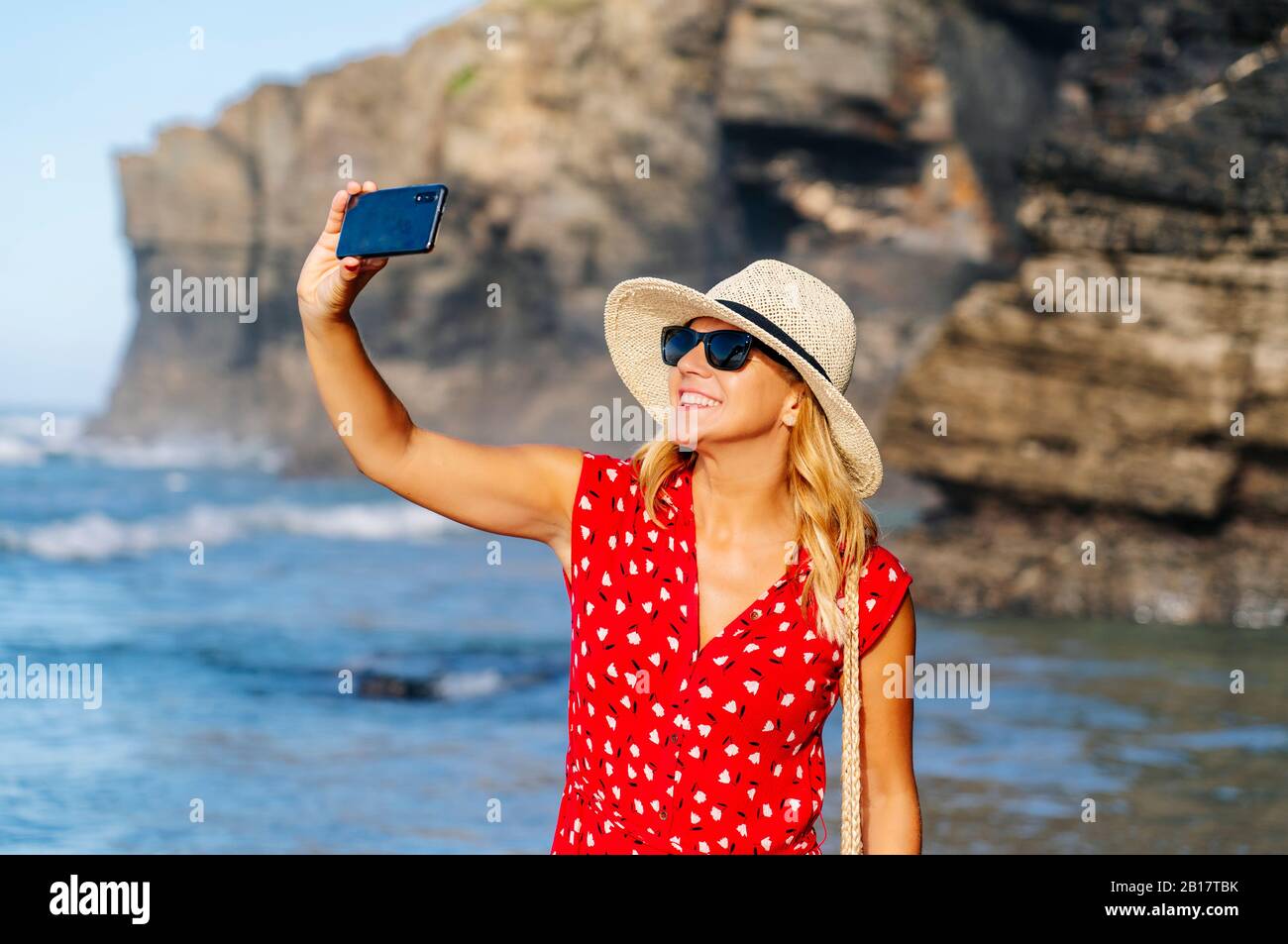 Blonde Frau in rotem Kleid und Hut und mit Smartphone und ein Selfie am Strand Stockfoto