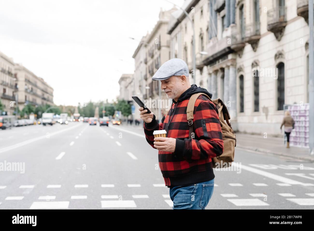 Älterer Mann mit Rucksack und Kaffee zum gehen, um auf Handy zu schauen, während die Straße überquert, Barcelona, Spanien Stockfoto