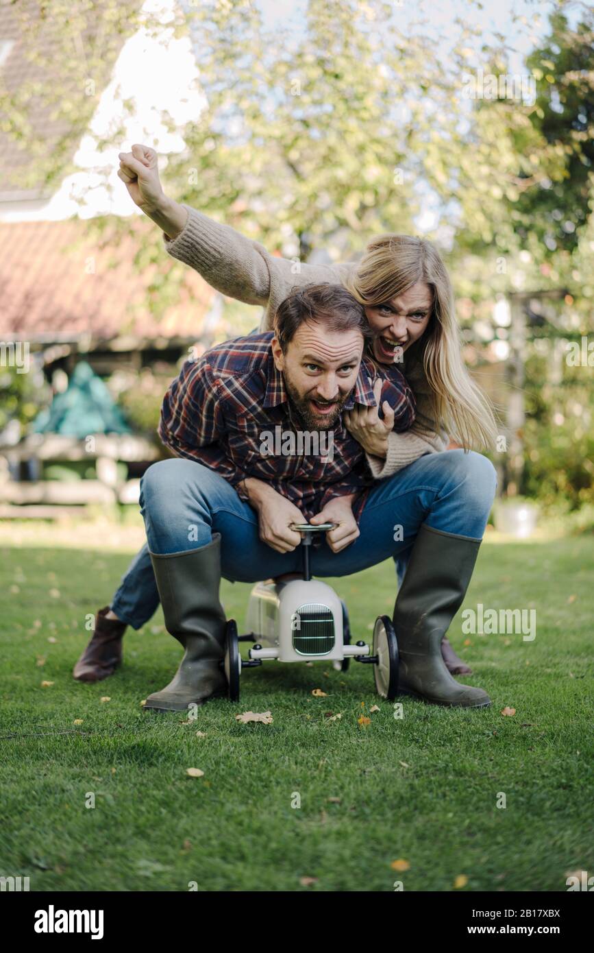 Ein lachendes Paar, das vorgibt, ein Spielzeugauto im Garten zu fahren Stockfoto