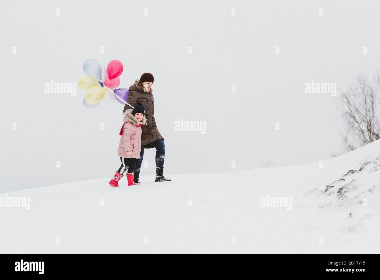 Mutter und kleines Mädchen mit Luftballons, die in schneebedeckter Landschaft laufen Stockfoto