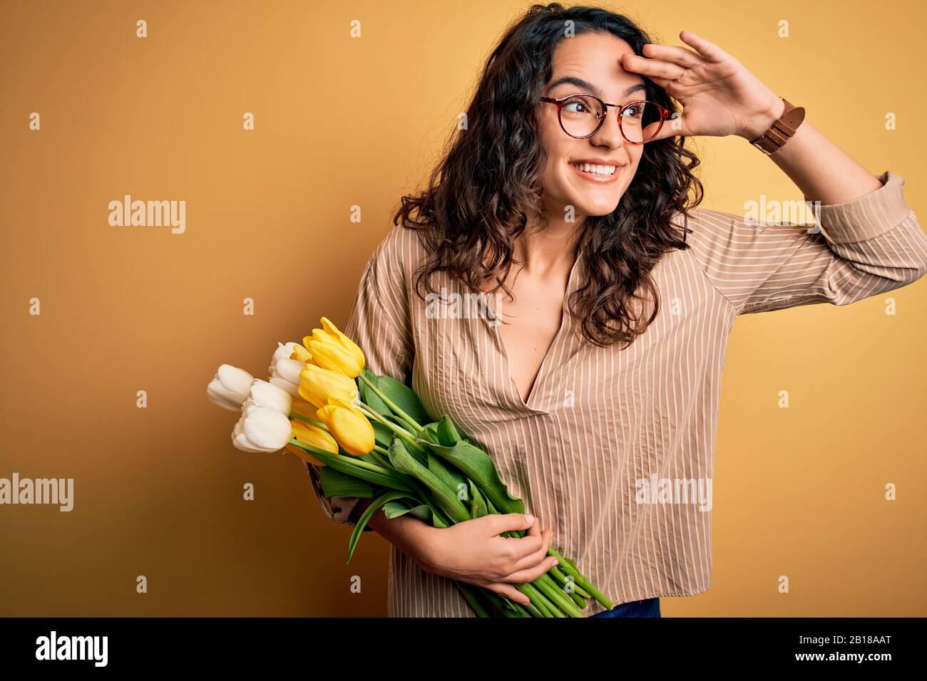 Junge, schöne romantische Frau mit lockigen Haaren, die einen Blumenstrauß mit gelben Tulpen hält, sehr glücklich und lächelnd, weit weg mit der Hand über den Kopf blickend. Suchen Stockfoto