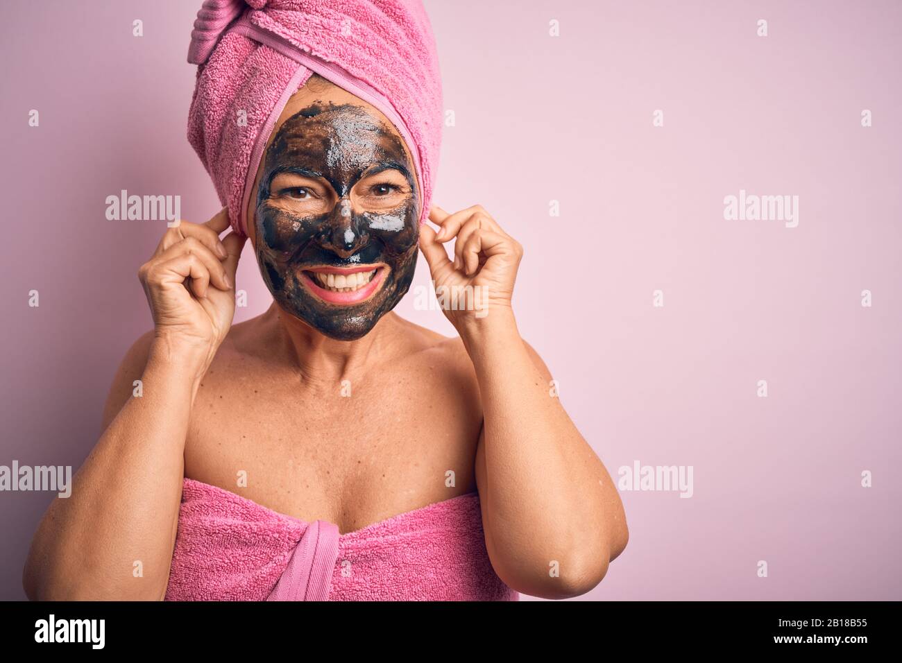 Frau im mittleren Alter, die schöne schwarze Gesichtsmaske über isoliertem rosafarbenem Hintergrund Trägt, Lächelnde Ohren mit Fingern ziehen, lustige Geste. Hörprobe Stockfoto