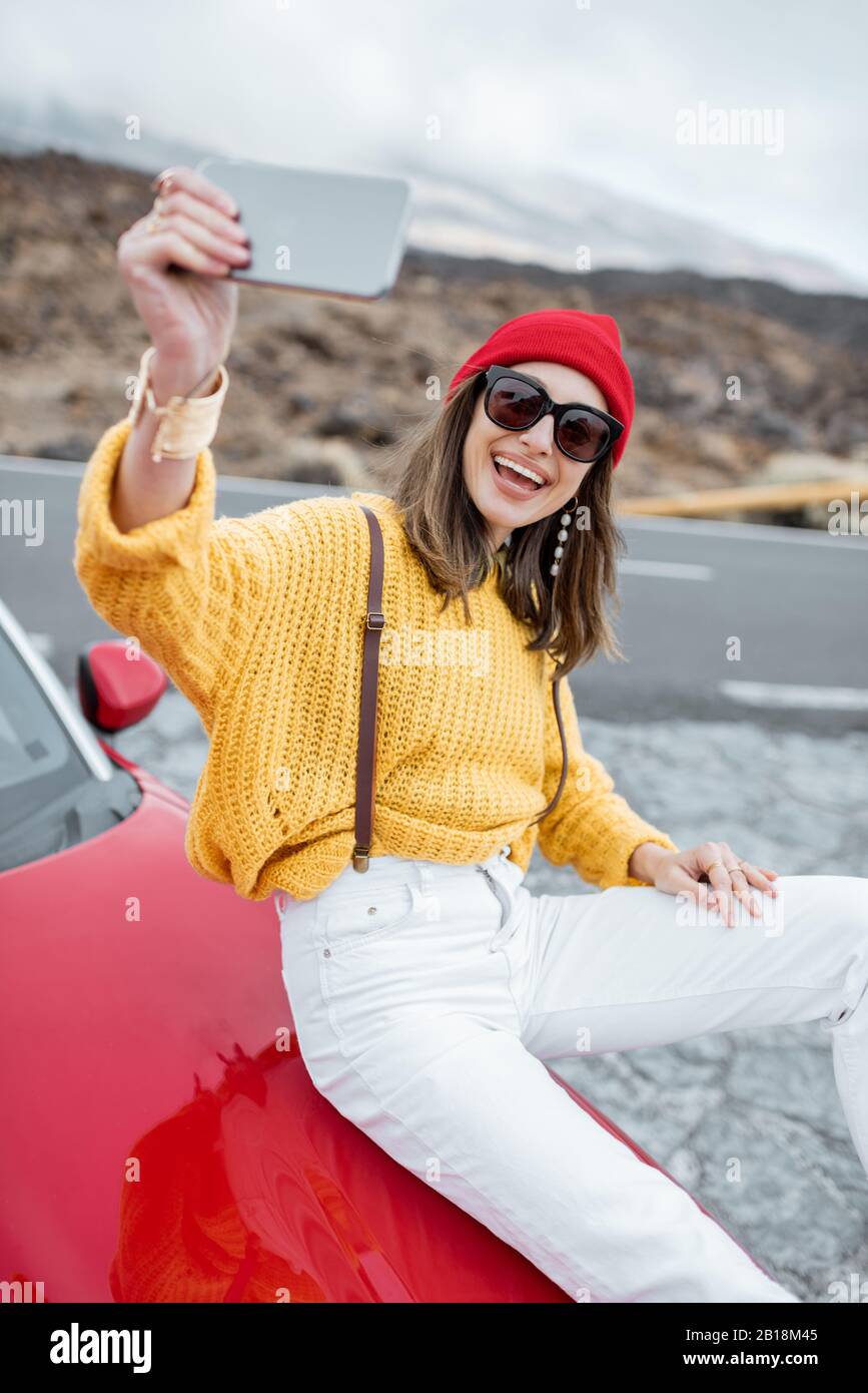 Junge Frau in hellem Pullover und Hut, die eine Fahrt auf der Straße im Wüstental genießen und auf dem Telefon fotografieren, während sie auf der Autohaube sitzt Stockfoto