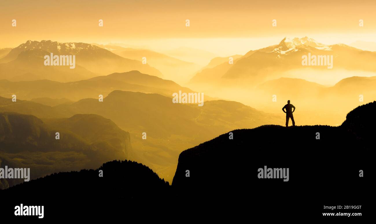 Spektakuläre Silhouetten der Bergketten. Mann erreicht Gipfel und genießt Freiheit. Sonnenaufgang mit orangefarbenem Licht. Stockfoto