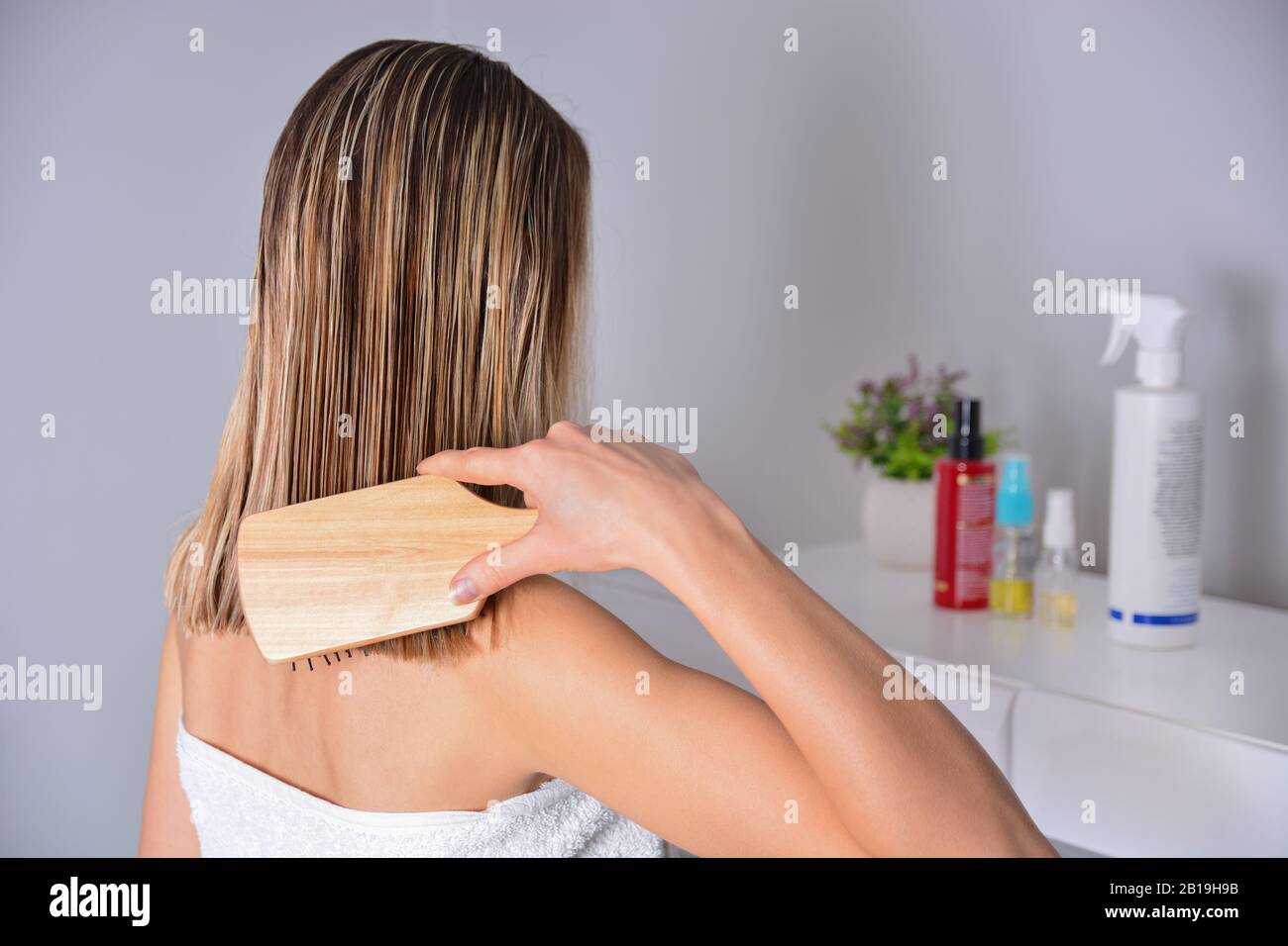 Frau mit Kammaufsatz, die ihr nasses blondes Haar nach dem Duschen zu Hause putzt. Sorgt für gesundes und sauberes Haar. Beauty-Salon-Konzept. Mädchen trägt weißes Handtuch Stockfoto