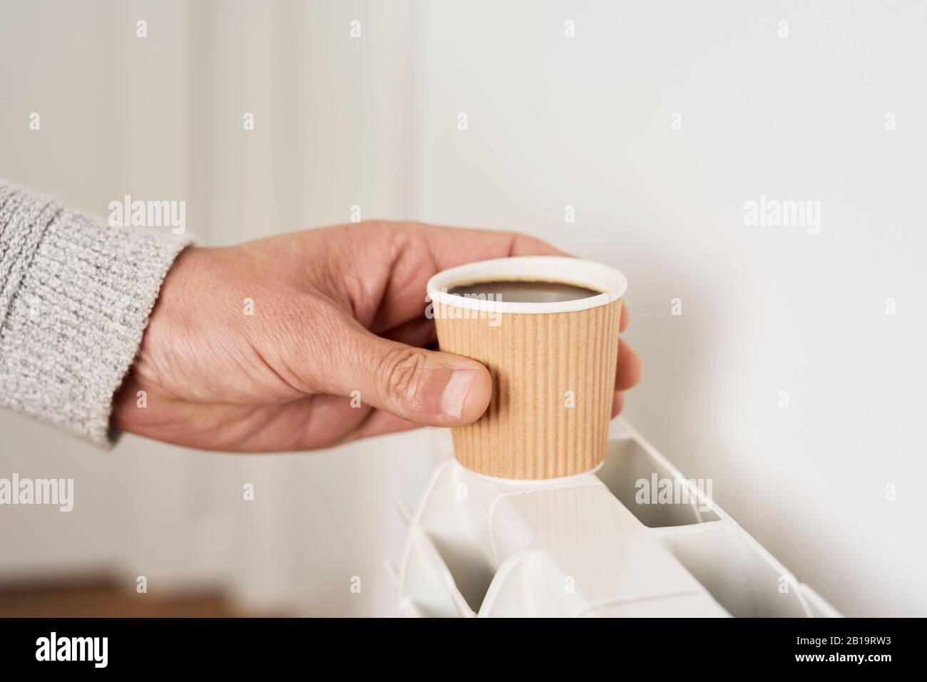 Nahaufnahme eines kaukasischen Mannes, der einen blassgrauen Pullover trägt und einen Papierbecher Kaffee auf einen Warmwasserkühler legt, um ihn heiß zu halten Stockfoto