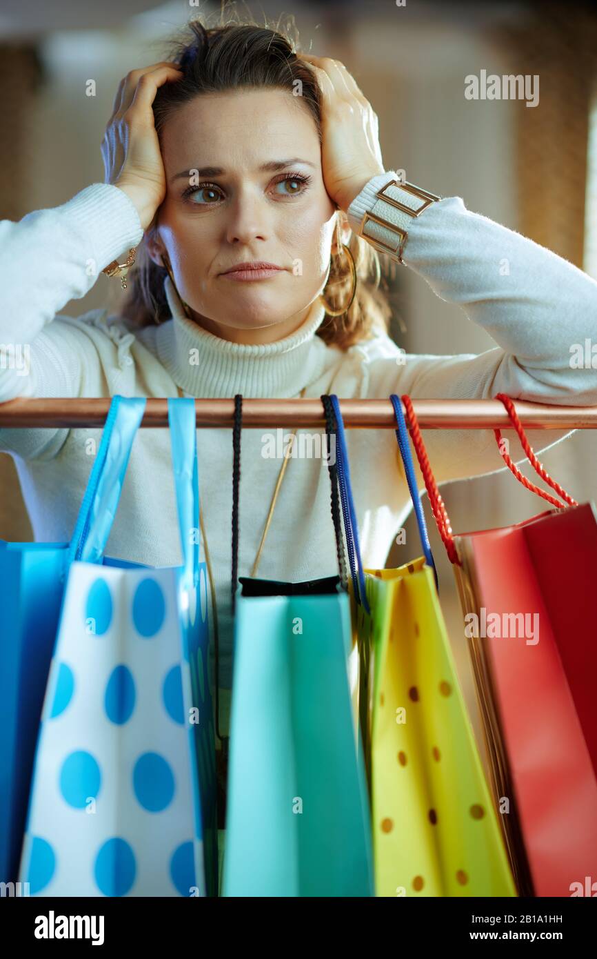 Verunglückte trendige Frau im mittleren Alter in weißem Pullover und Rock in der Nähe von bunten Einkaufstaschen, die an der Kupfer-Kleiderschiene hängen. Stockfoto