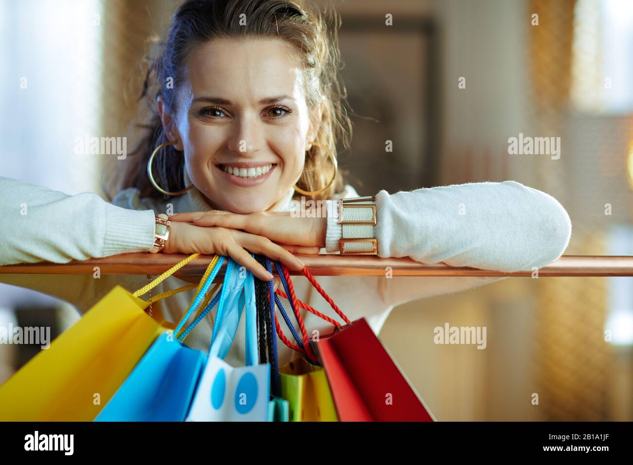 Porträt der lächelnden eleganten Frau in weißem Pullover und Rock in der Nähe von bunten Einkaufstaschen hängen an der Kupfer-Kleiderschiene. Stockfoto