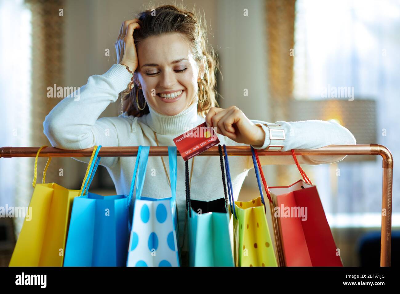 Lächelnde, stilvolle Frau in weißem Pullover und Rock mit Kreditkarte in der Nähe von bunten Einkaufstaschen, die an der Kupfer-Kleiderschiene hängen. Stockfoto