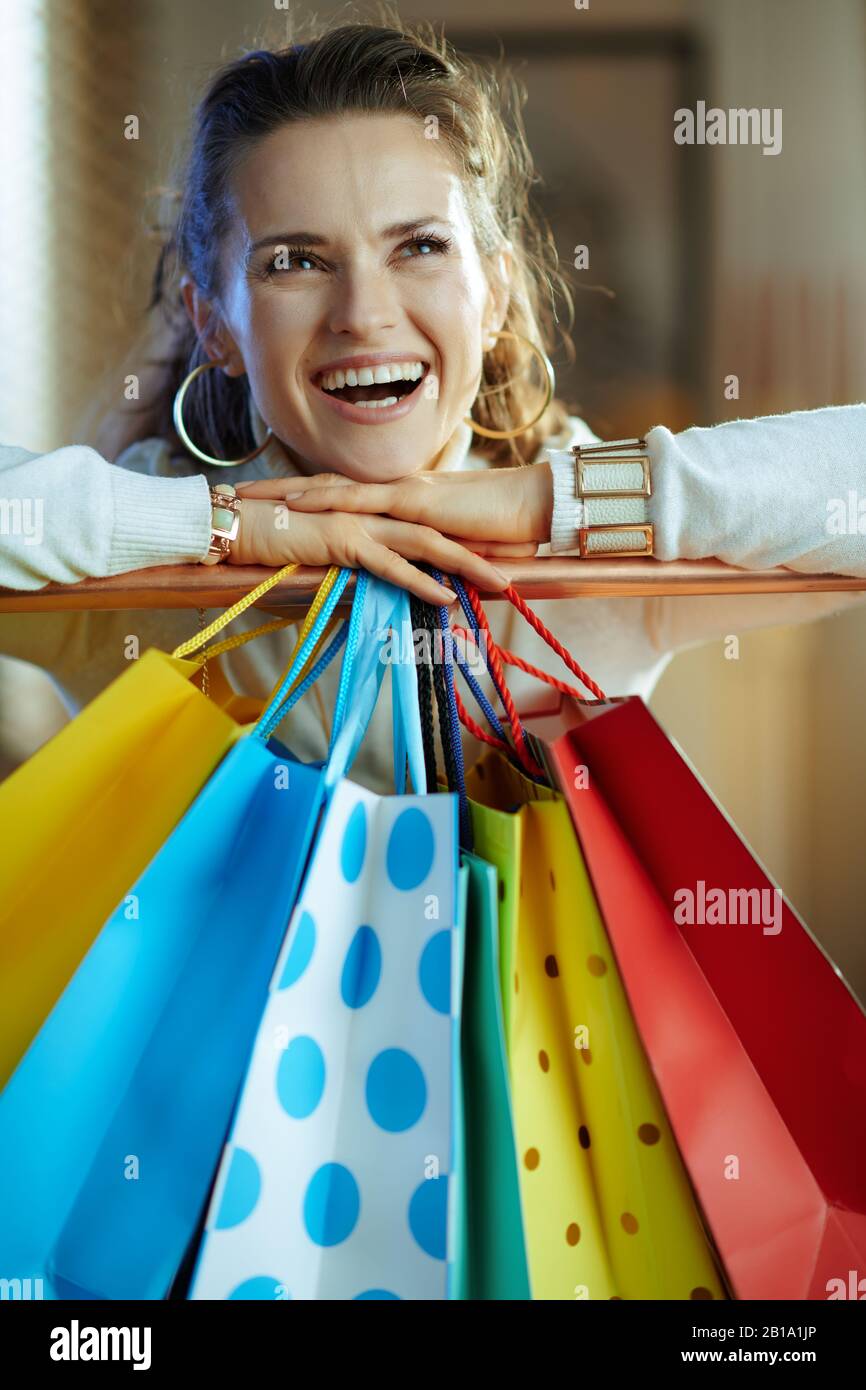 Lächelnde, elegante 40 Jahre alte Frau in weißem Pullover und Rock, die in die Ferne blickt, in der Nähe von bunten Einkaufstaschen, die an der Kupfer-Kleiderschiene hängen. Stockfoto