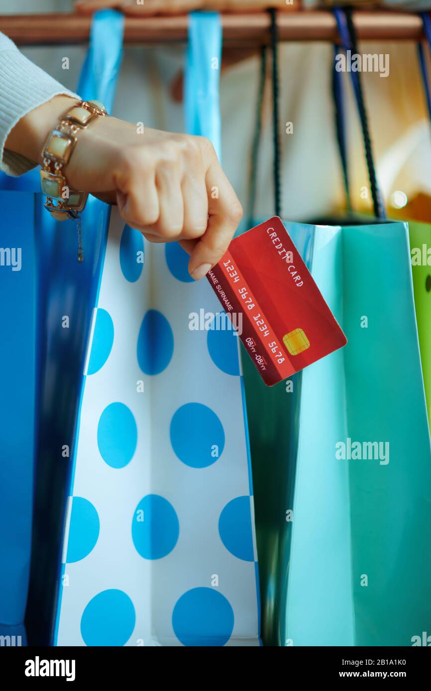 Nahaufnahme der jungen Frau in weißem Pullover und Rock mit Kreditkarte in der Nähe von bunten Einkaufstaschen, die an der Kupfer-Kleiderschiene hängen. Stockfoto