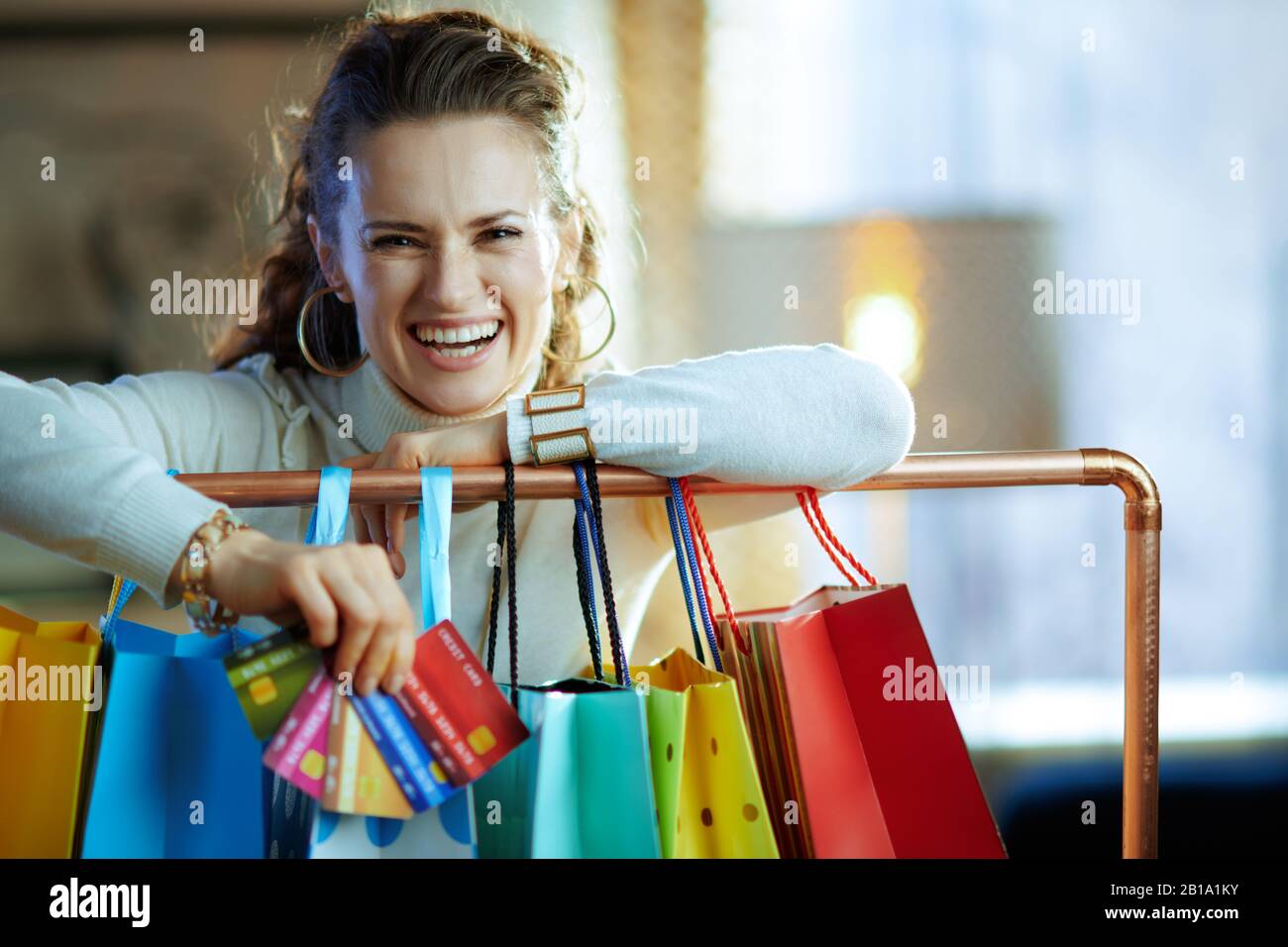 Porträt einer lächelnden, stylischen 40 Jahre alten Frau in weißem Pullover und Rock in der Nähe farbenfroher Einkaufstaschen, die an der Kupfer-Kleiderschiene mit einem Fan von Cre hängen Stockfoto