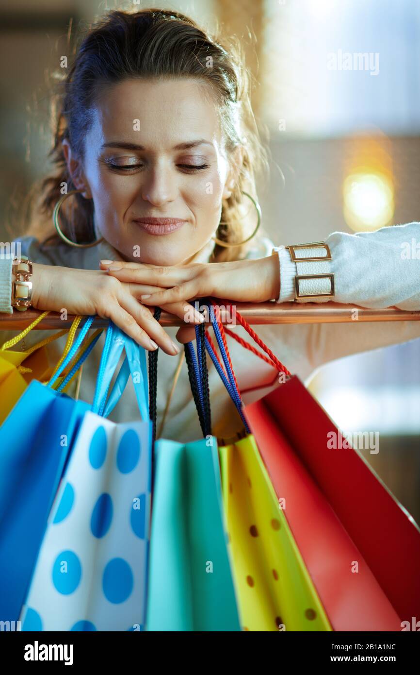 Porträt der 40-jährigen entspannten Frau in weißem Pullover und Rock in der Nähe von bunten Einkaufstaschen, die an der Kupfer-Kleiderschiene hängen. Stockfoto