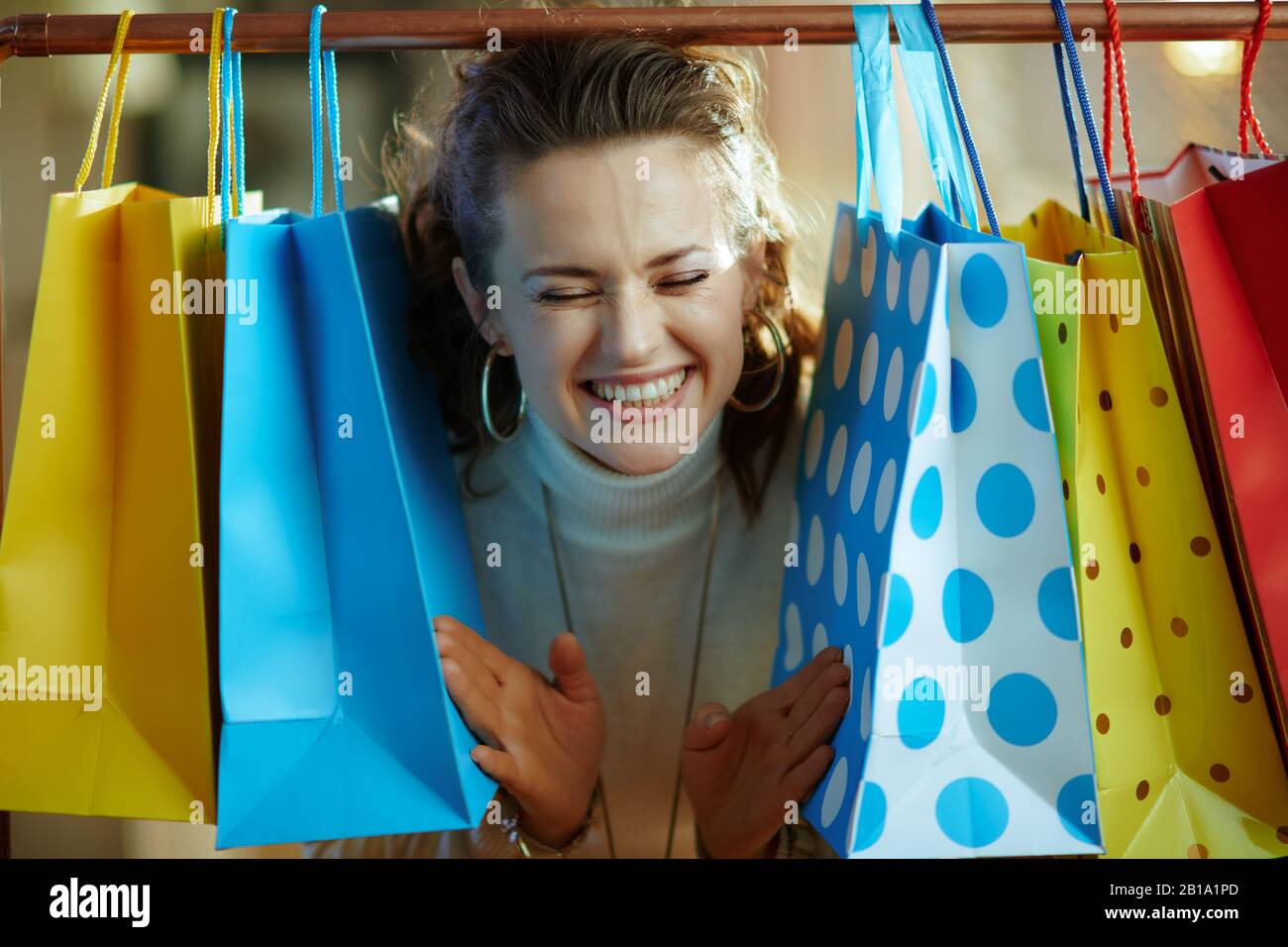 Fröhliche, moderne Frau in weißem Pullover und Rock, die aus bunten Einkaufstaschen, die an der Kupfer-Kleiderschiene hängen, herausticht. Stockfoto