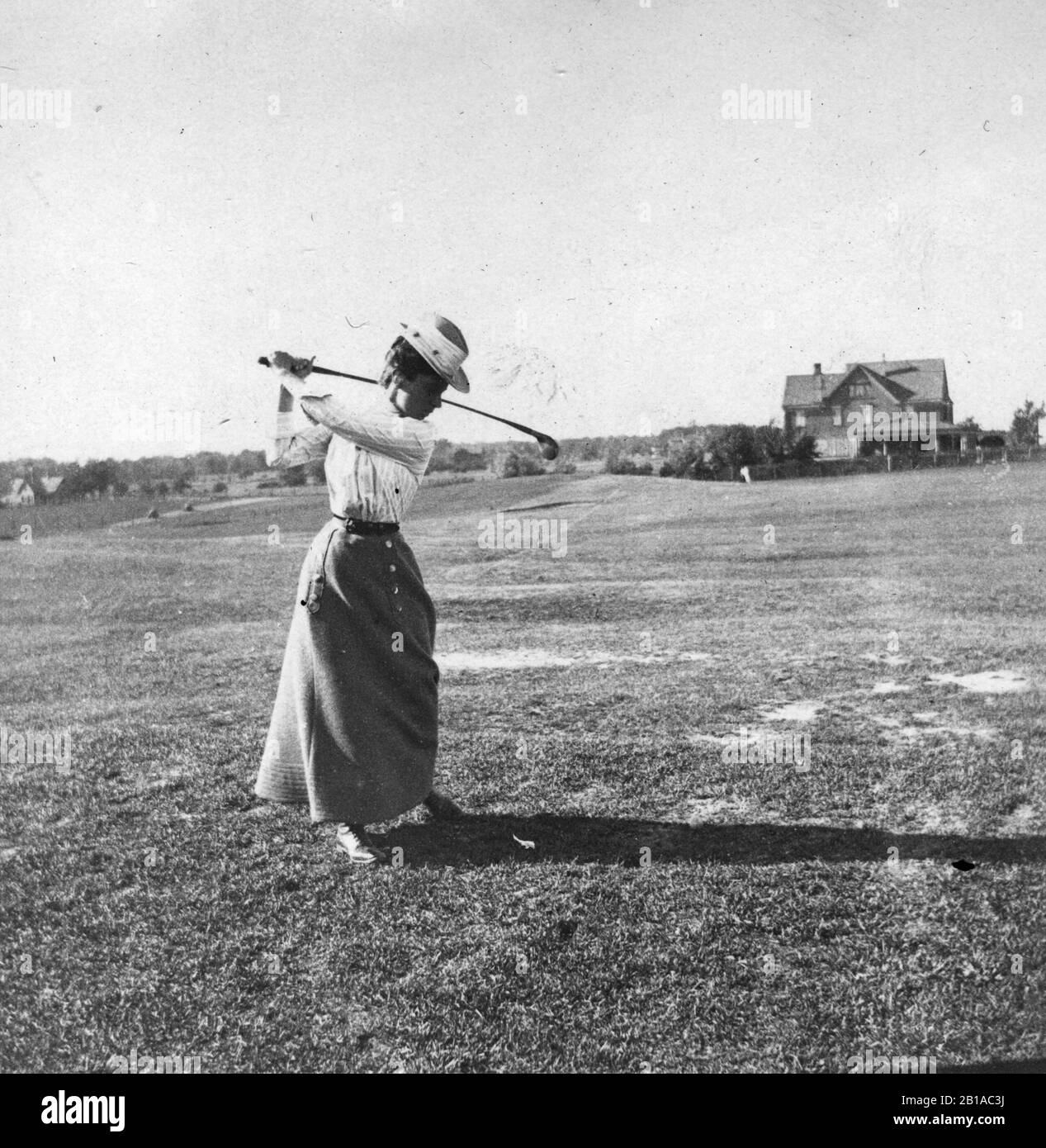 Frau im Golf-Backswing, um ihren Ball auf dem Platz mit ihrem Holz (kein Eisen) zu schlagen. c. 1910. Der Golfplatz ist ein leicht rollendes, beschnittenes Feld. Die Dame ist modisch angezogen, obwohl ihr Rock knöchellang ist. Der Ausweis der Dame ist unbekannt. Um meine anderen Golf-bezogenen Vintage-Bilder zu sehen, suchen Sie: Prestor vintage Sport Stockfoto