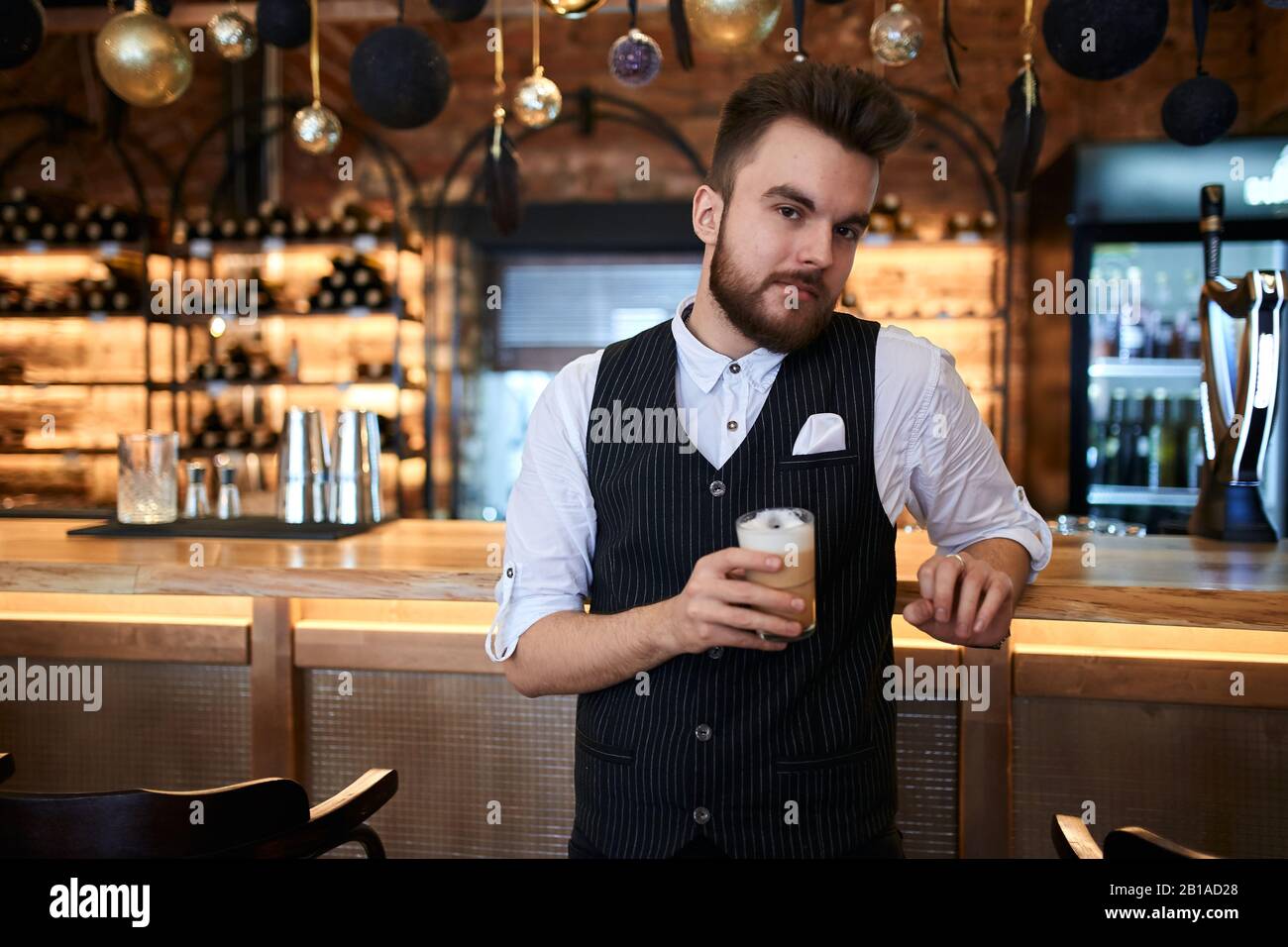 Gutaussehender junger bärtiger Mann, der die Kaffeetasse hält und die Kamera ansieht, während er an der Theke steht, Nahaufnahme Stockfoto