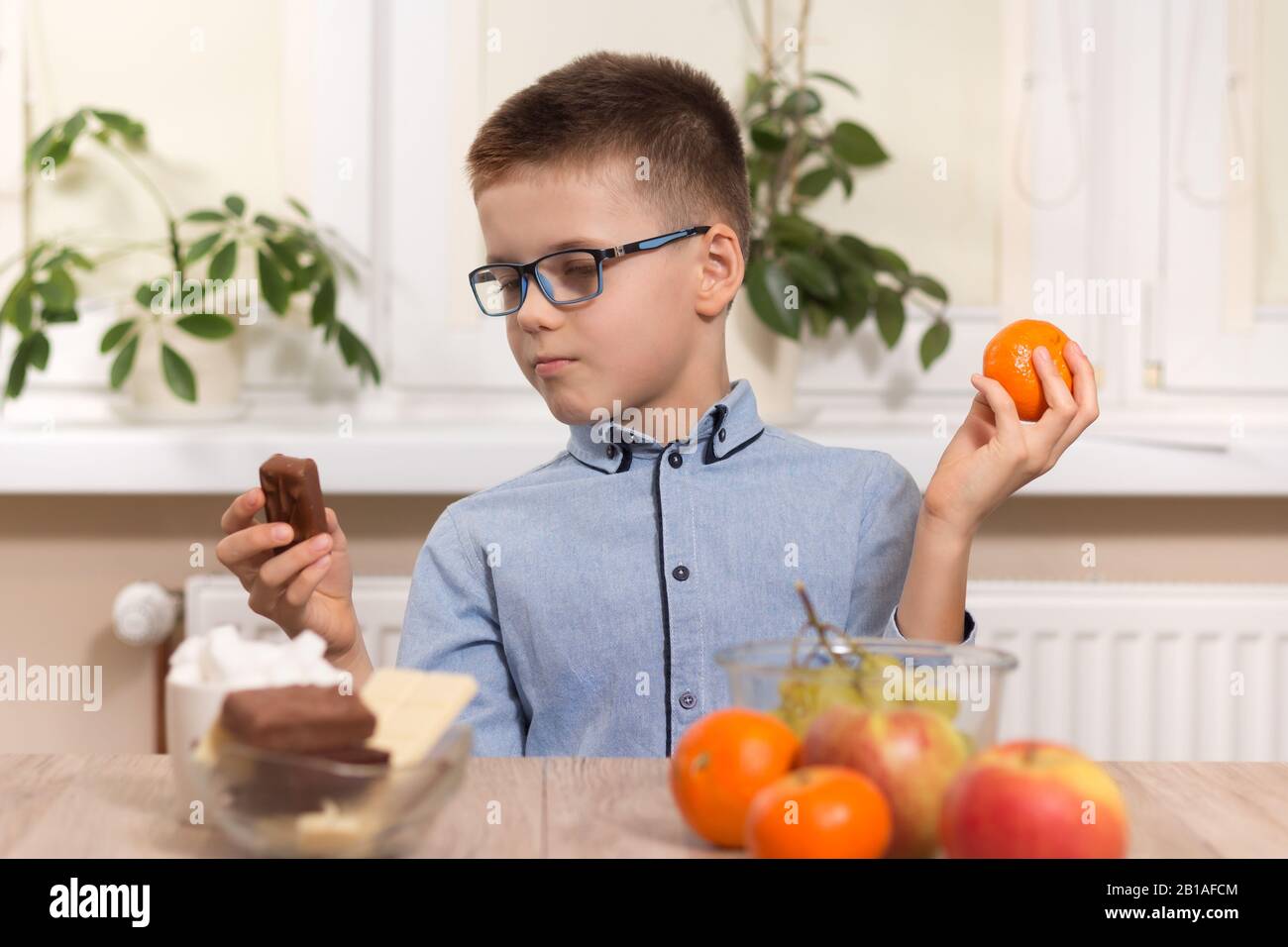 Schokoladenriegel oder Tangerinfrucht. Der schulische Junge sitzt am Tisch und fragt sich, was er wählen soll. Stockfoto