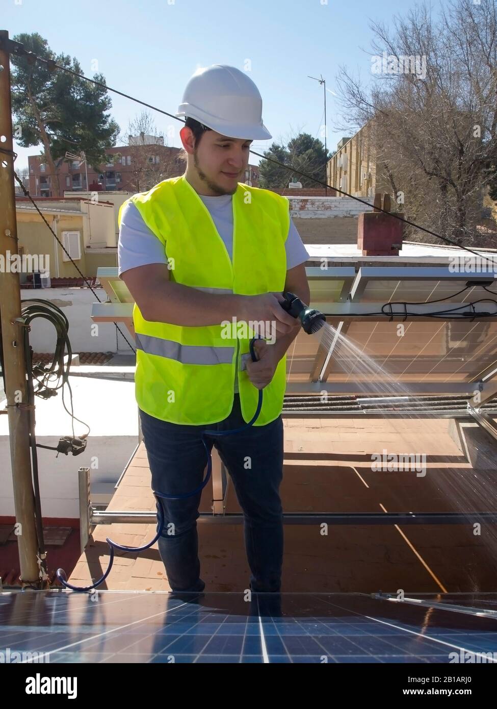 Kaukasisch attraktiver junger Techniker, der Solarpaneele mit einem Schlauch reinigt. Alternative Stromquelle, nachhaltige Ressourcenkonzeption. Stockfoto