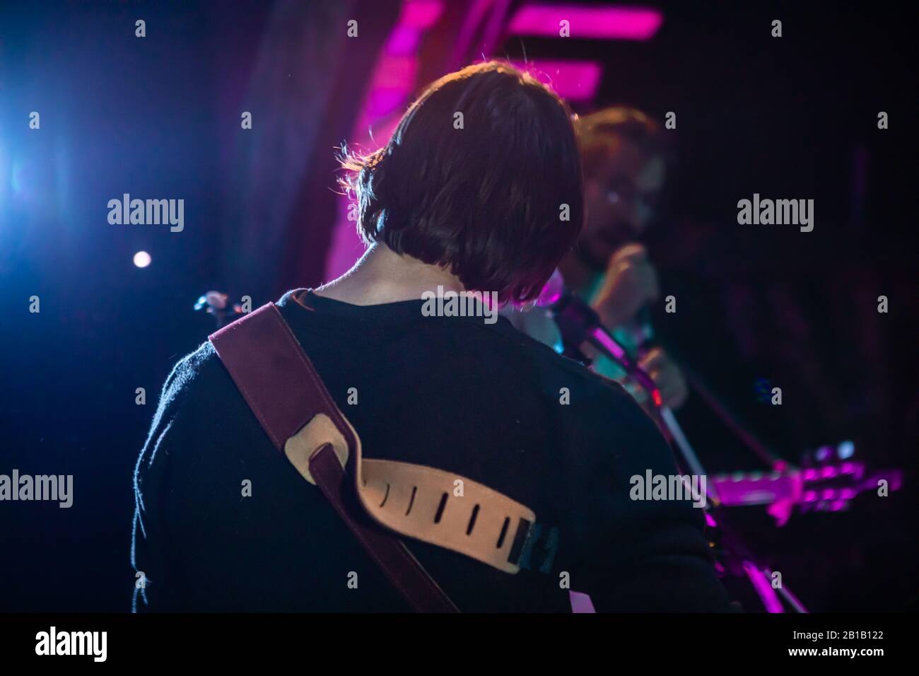 Eine Nahaufnahme von hinter einem Musiker, der auf der Bühne mit verschwommenen Bandmitgliedern, im Hintergrund und violettem Bühnenlicht bei Nacht auf dem Festival auftrat Stockfoto