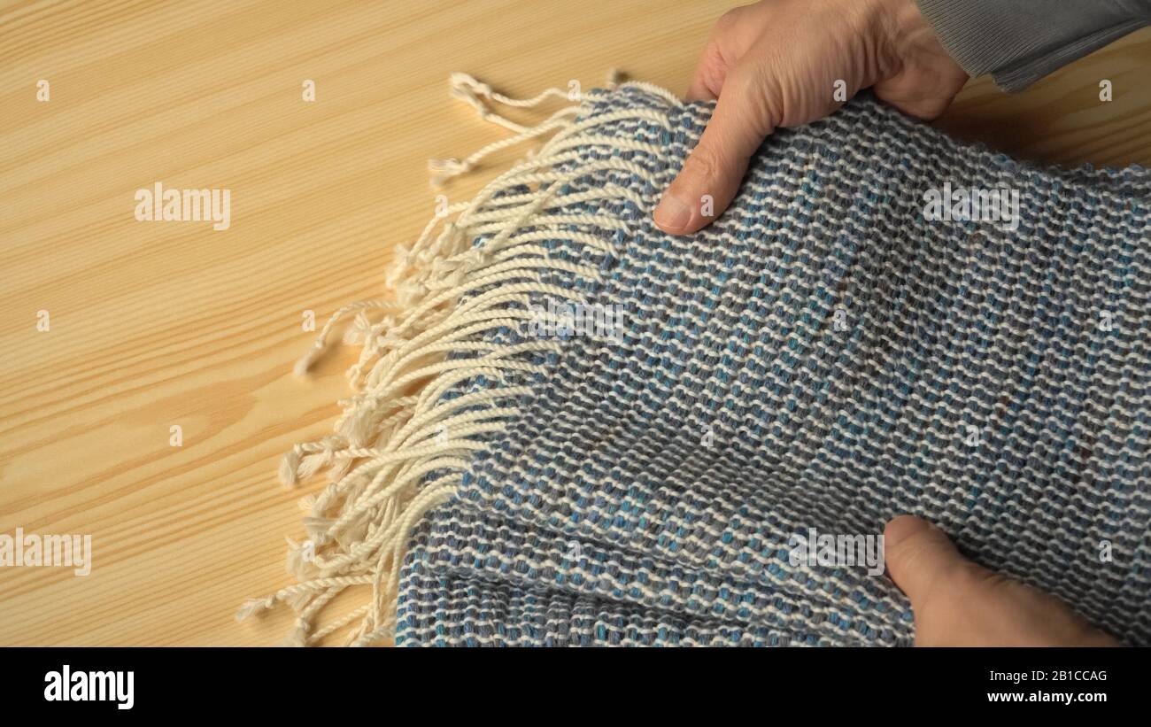 Handgefertigter Wollschal in den Händen des Strickers. Handgewebte Waren aus Webstühlen. Eine Nahansicht zeigt weiß gestreiftes gewebtes Gewebe mit dunkleren Bändern in Blau-GREE Stockfoto