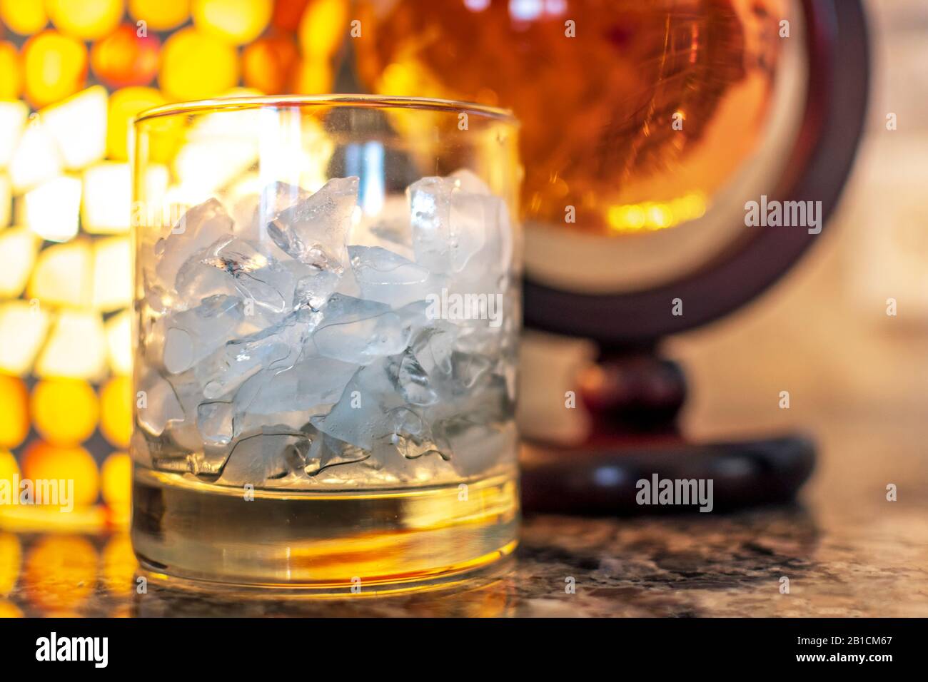 Ein Alkoholglas aus zerkleinertem Eis sitzt neben einem runden Behälter mit Scotch-Whisky auf einem Granit mit warmem Licht Stockfoto