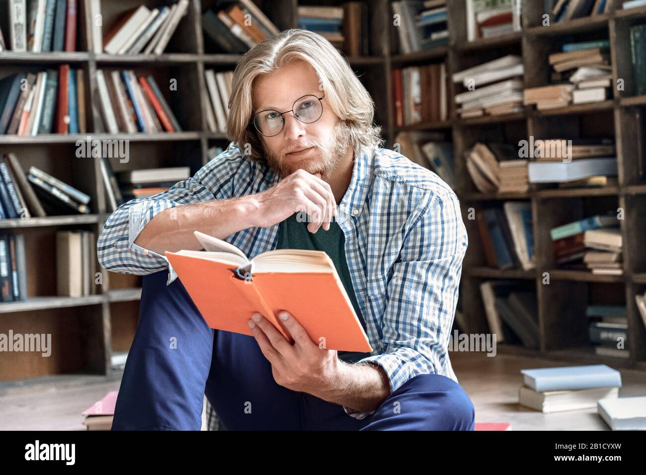 Nachdenkliche junge männliche Studentenbrille las das Boden-Bildungskonzept der Buchbibliothek Stockfoto