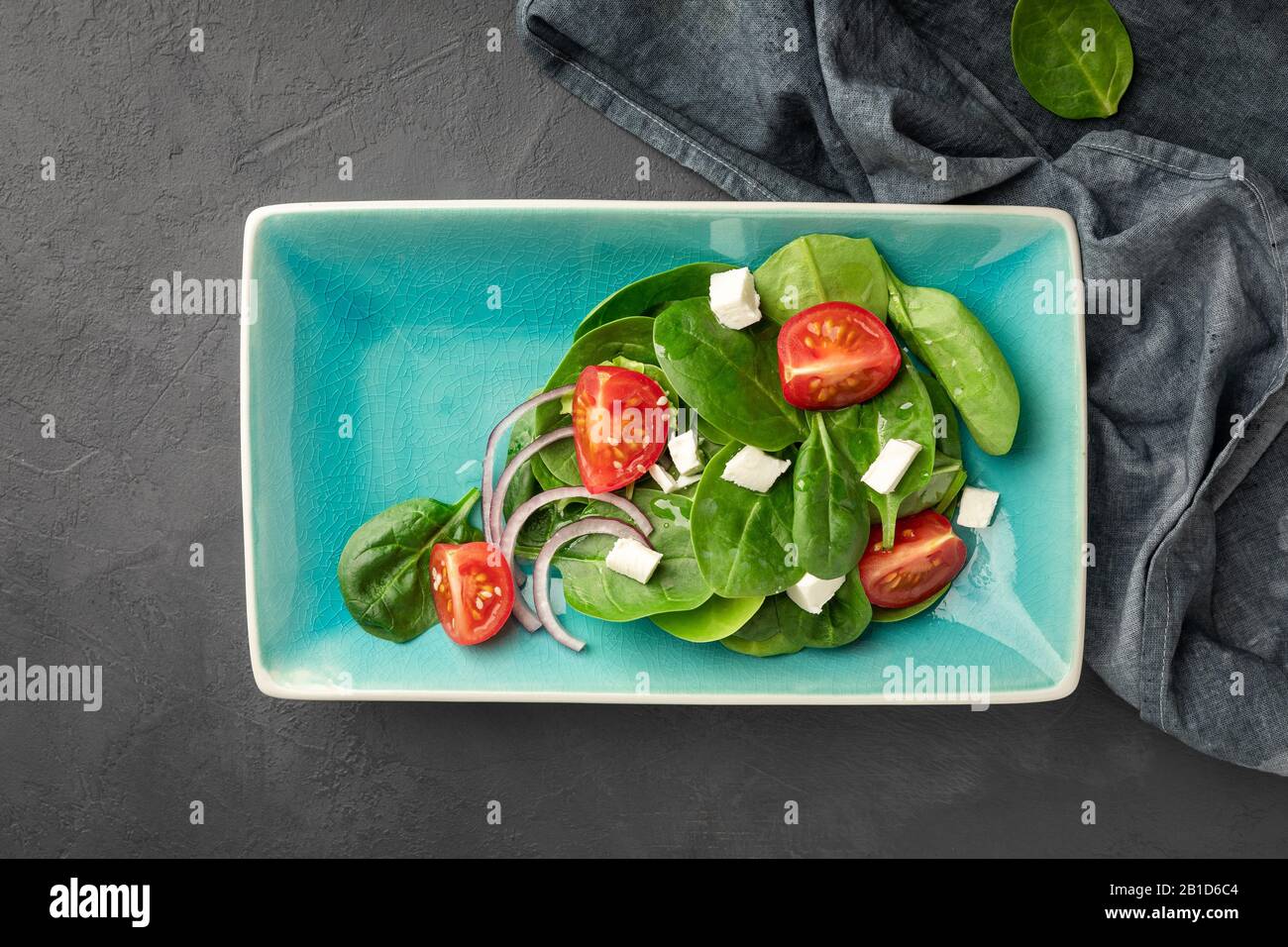 Blick auf einen frischen grünen Gemüsesalat aus Spinat, Tomaten und Sesamsamen auf einer blauen, rechteckigen Platte. Schwarzer Hintergrund. Stockfoto