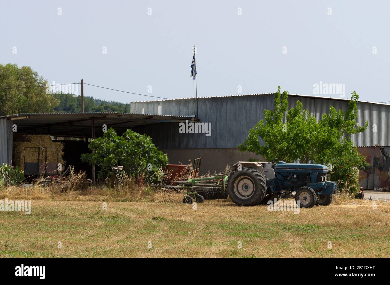 Blauer Traktor im Nahbereich auf einem gemähten Feld vor dem Hintergrund eines Gebäudes mit einer herabhängenden griechischen Flagge (Rhodos, Griechenland) Stockfoto