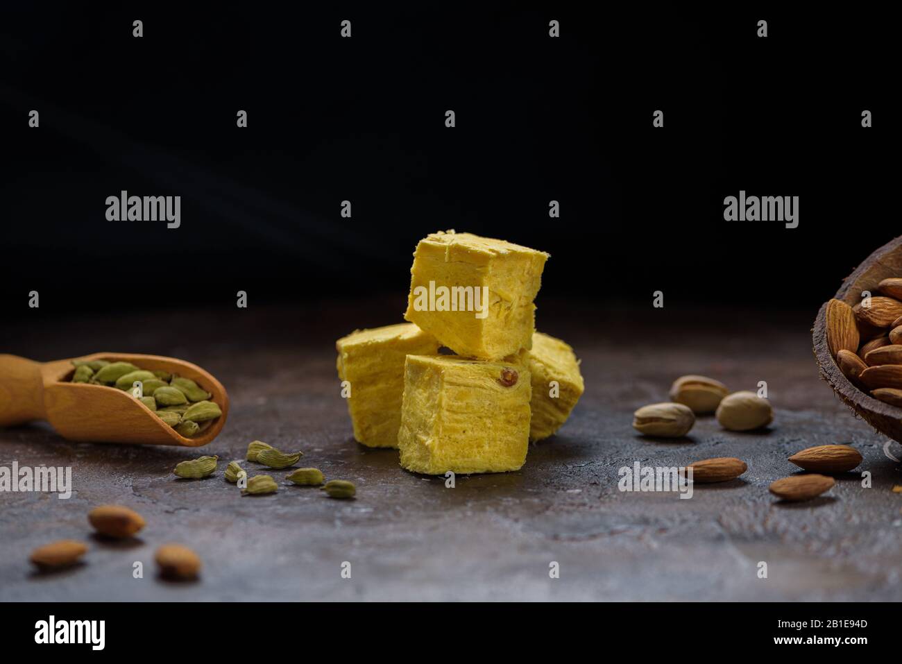 Würfel von Dessert soan Papdi, Kardamom Körner in Holzlöffel, Pistazien und Mandel auf Beton Küchenoberfläche mit schwarzem Kopieplatz. Stockfoto