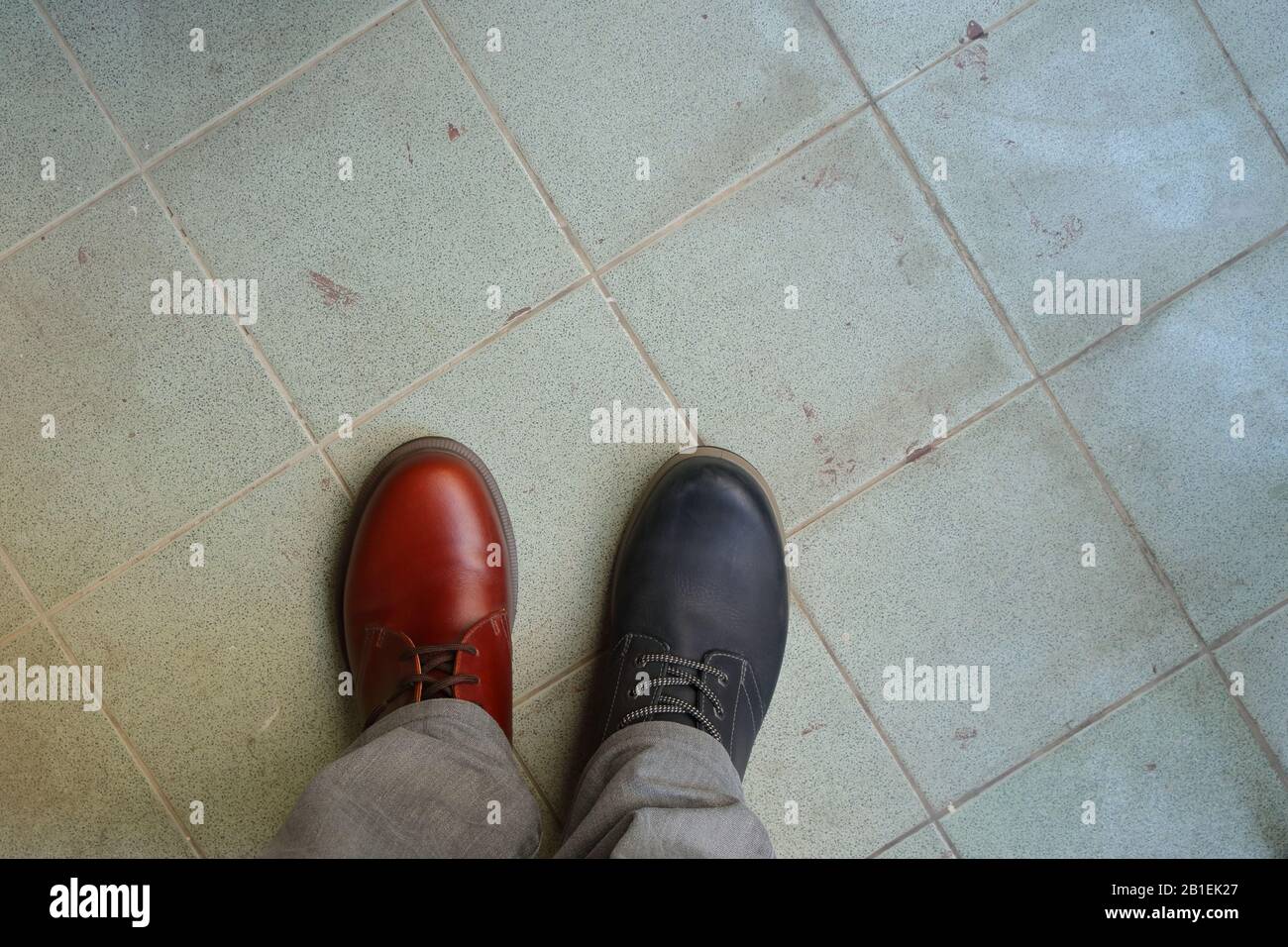 Das Obermaterial aus zusammenpassenden Schuhen, ein Mann, der zwei verschiedene Schuhe und verschiedene Farben auf gefliestem Boden trägt, bricht die Regeln, revolutioniert die Metaphik Stockfoto