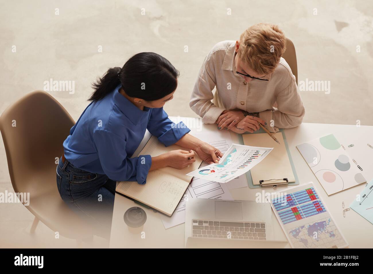 Hochwinkelansicht bei zwei modernen Geschäftsfrauen, die Datendiagramme diskutieren, während sie am Tisch sitzen und Platz für Kopien schaffen Stockfoto