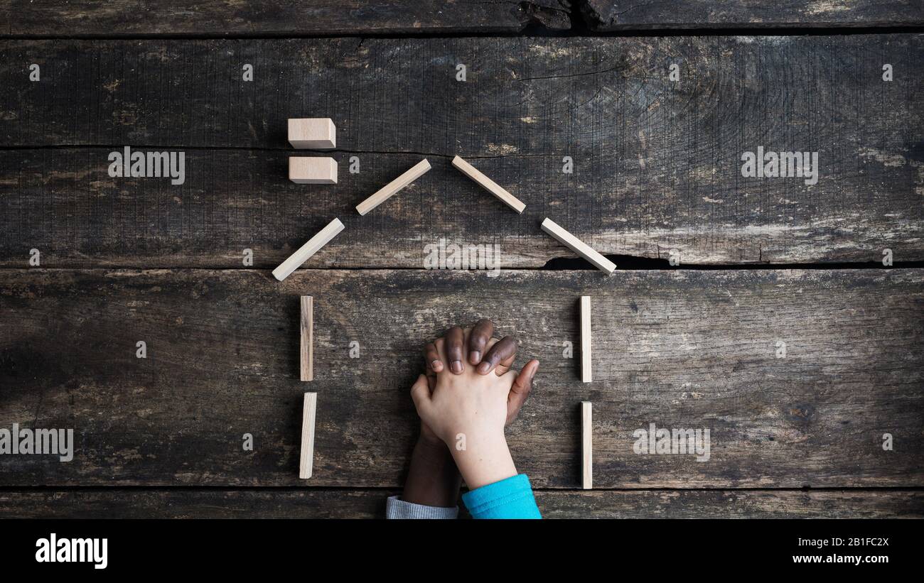 Gleichberechtigung und Frieden konzeptionelles Bild - Hand von kaukasischen und farbigen Kindern, die sich in einem Haus aus Ooden halten. Über Holzhintergrund Stockfoto