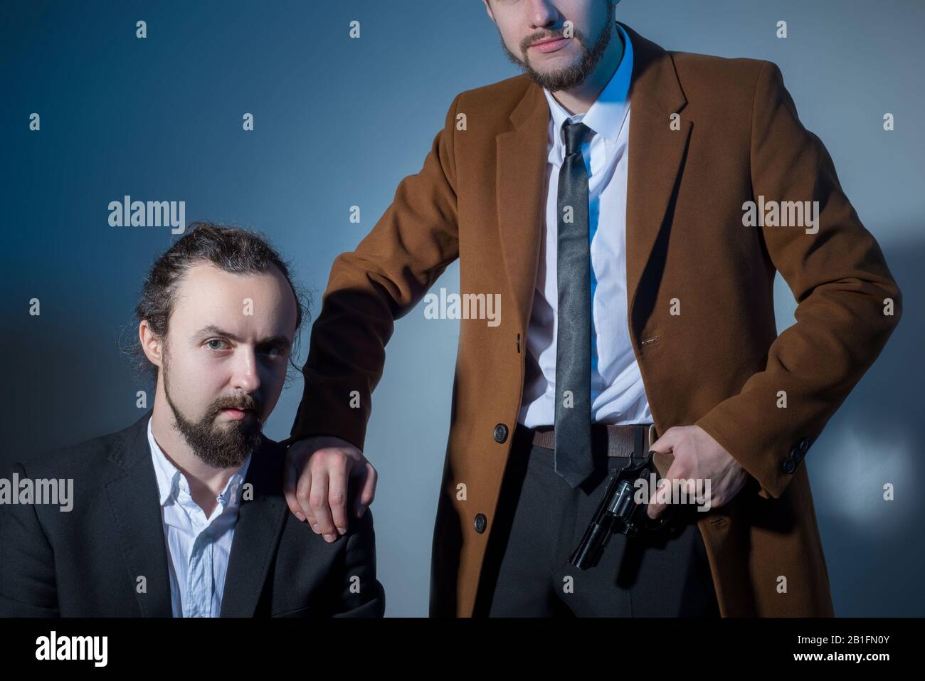 Porträt von zwei Männer in Anzügen, einer von Ihnen sitzt, die andere im Besitz einer Waffe. Auf grauem Hintergrund. Dramatische Licht. Künstlerische Einstellung. Die Stockfoto