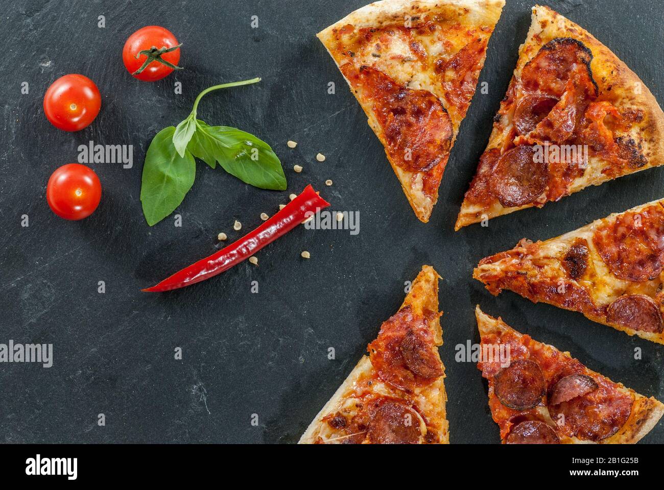 Köstliche frisch gekochte Scheiben von Pfefferoni Pizza auf Schiefer mit Kirschtomaten, rotem Pfeffer und Basilikumblättern Stockfoto