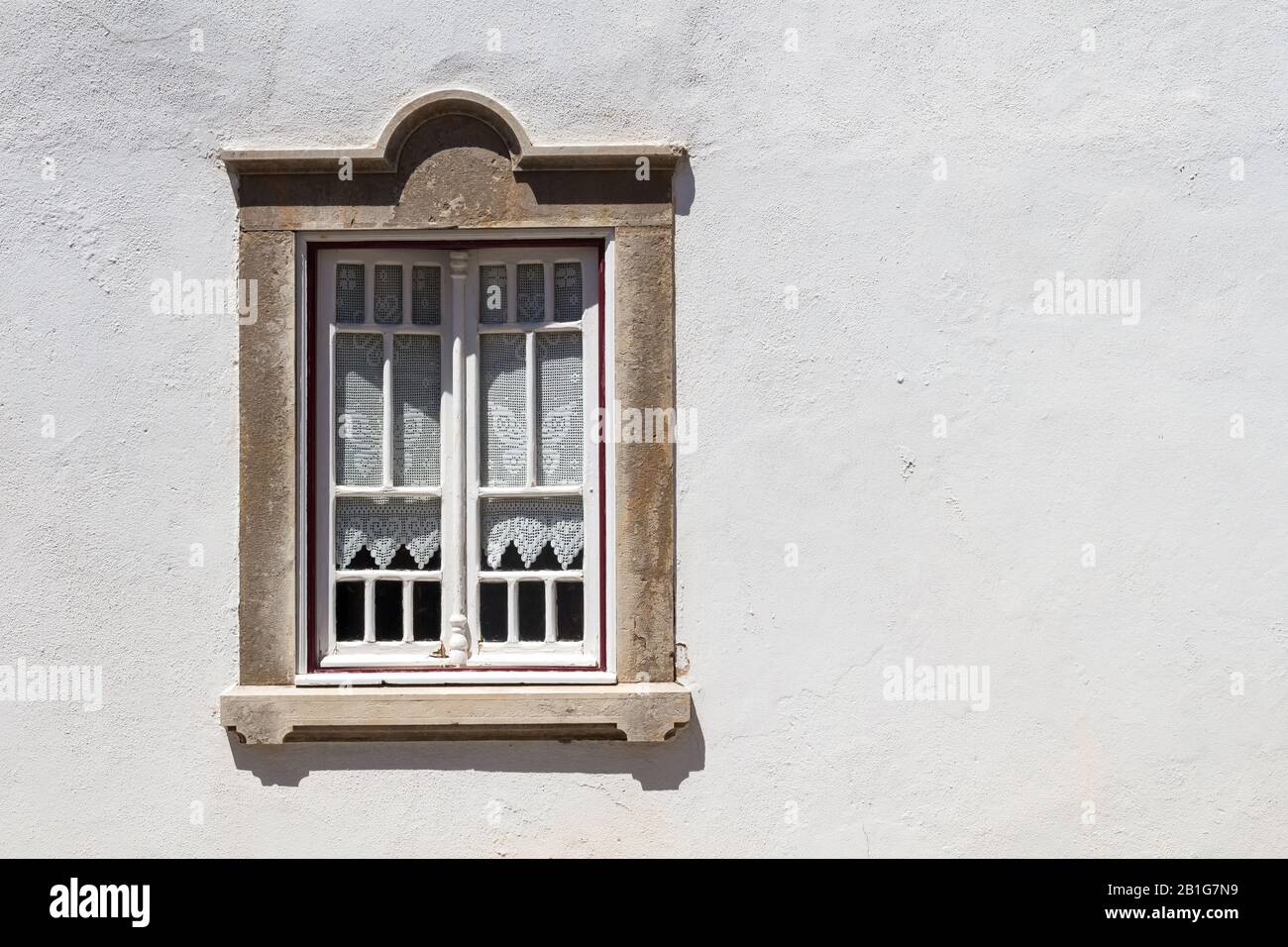 Helle weiße Hauswand an einem sonnigen Tag. Fenster mit weißen Holzdetails und altmodischen Vorhängen. Steinrahmen. Estoi, Portugal. Stockfoto