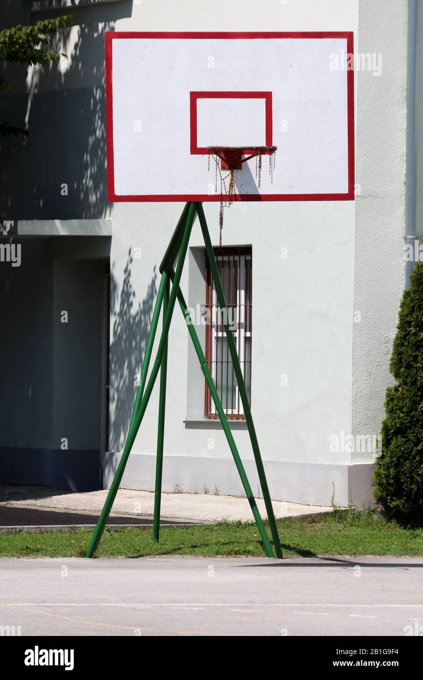 Renovierter und umlackierter Basketballkorb mit einem alten verrosteten, gebrochenen Kettennetz, das auf einem starken Metallrahmen am Rande des gepflasterten Basketballplatzes der High School montiert ist Stockfoto