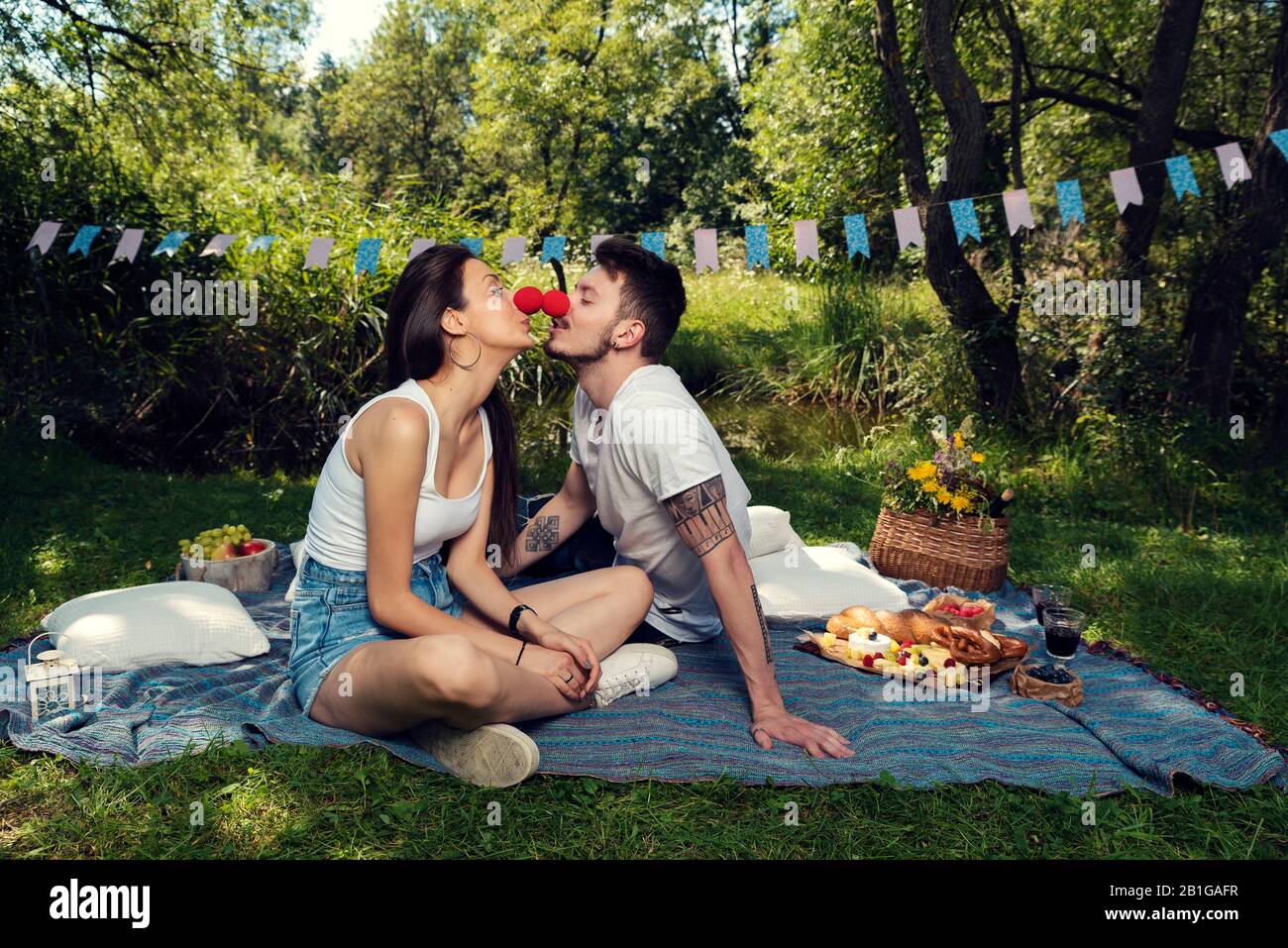 Junges Paar bei einem Picknick in einem Stadtpark, das auf einer Decke sitzt, die rote Nasen trägt und sich gegenseitig küsst Selective Focus Stockfoto