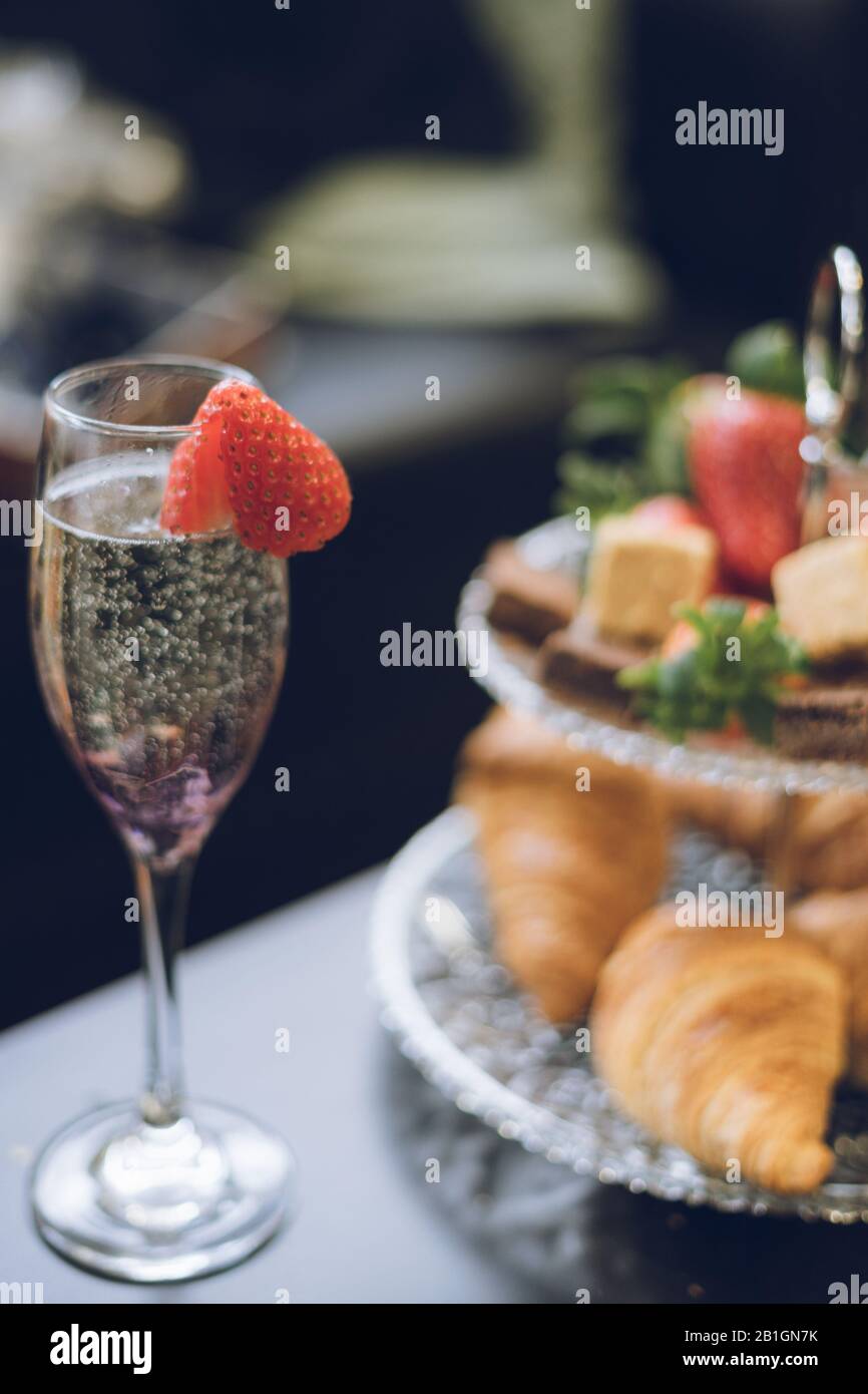 Eine Nahaufnahme einer Erdbeere auf einem Champagnerglas mit Croissants Erdbeeren und Kuchen auf einem gestuften Tortenständer im Hintergrund Stockfoto