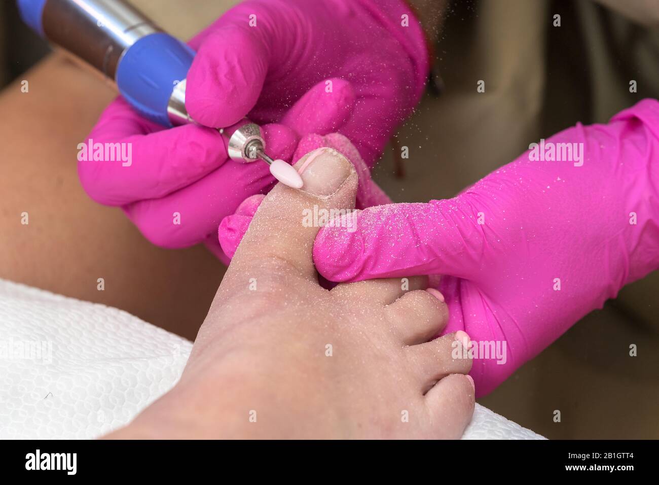Der Pediküre-Prozess. Die pedikuristische Frau entfernt mit einem elektrischen Nagelbohrer alte gelpoliermittel von den Nägeln auf Zehen. Weibliche Hände mit speziellem Gummipink Stockfoto