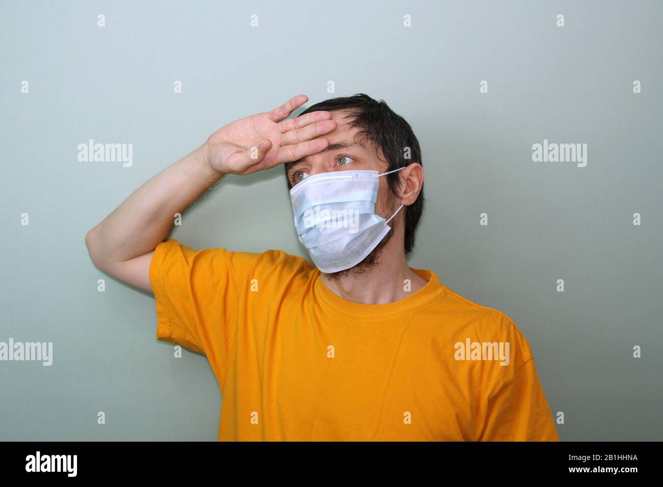 Erwachsene Männer mittleren Alters in einem gelben T-Shirt auf grauem Hintergrund in einer medizinischen Maske halten seine Hand nahe seiner Stirn. Kopfschmerzen, hohes Fieber. Krankheitsschutz Coronavirus epidemisches Problem aus China. Stockfoto