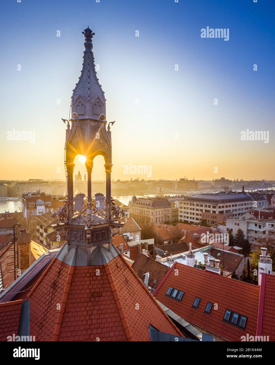 Budapest, Ungarn - Alter traditioneller Metallturm und rote Dächer des Burgviertels mit aufsteigender Sonne, St. Stephens-Basilika und Szechenyi-Kettenbrücke Stockfoto