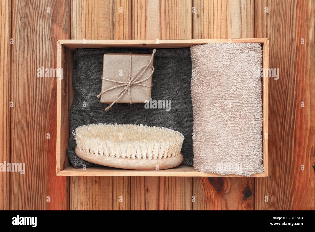Handgefertigte Seife, die in Handpapier, Handtuch und Massagebürste gewickelt ist, liegen in einer Holzkiste auf einem Holzhintergrund. Stockfoto