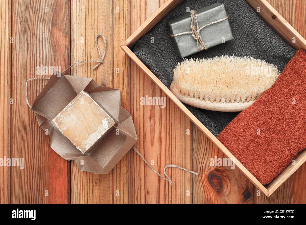 Handgefertigte Seife, die in Handpapier, Handtuch und Massagebürste gewickelt ist, liegen in einer Holzkiste auf einem Holzhintergrund. Stockfoto