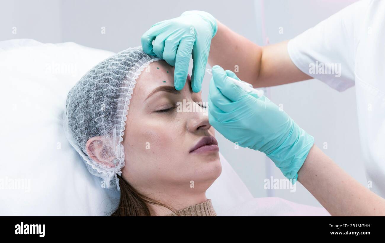 Die junge Kosmetikerin, die sich auf die Injektion in die weibliche Stirn vorbereitet. Der Arzt Kosmetologe macht Anti-Aging-Behandlung und Gesichtsaufhebung probed Stockfoto